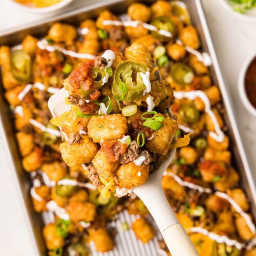 closeup of spatula containing tater tots with background of sheet pain with tater tots surrounded by bowls of cheese, onion, jalapeno and salsa