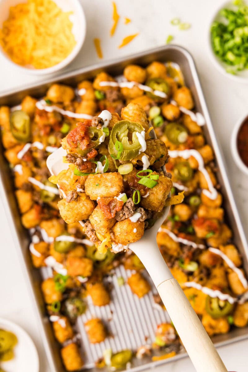 closeup of spatula containing tater tots with background of sheet pain with tater tots surrounded by bowls of cheese, onion, jalapeno and salsa