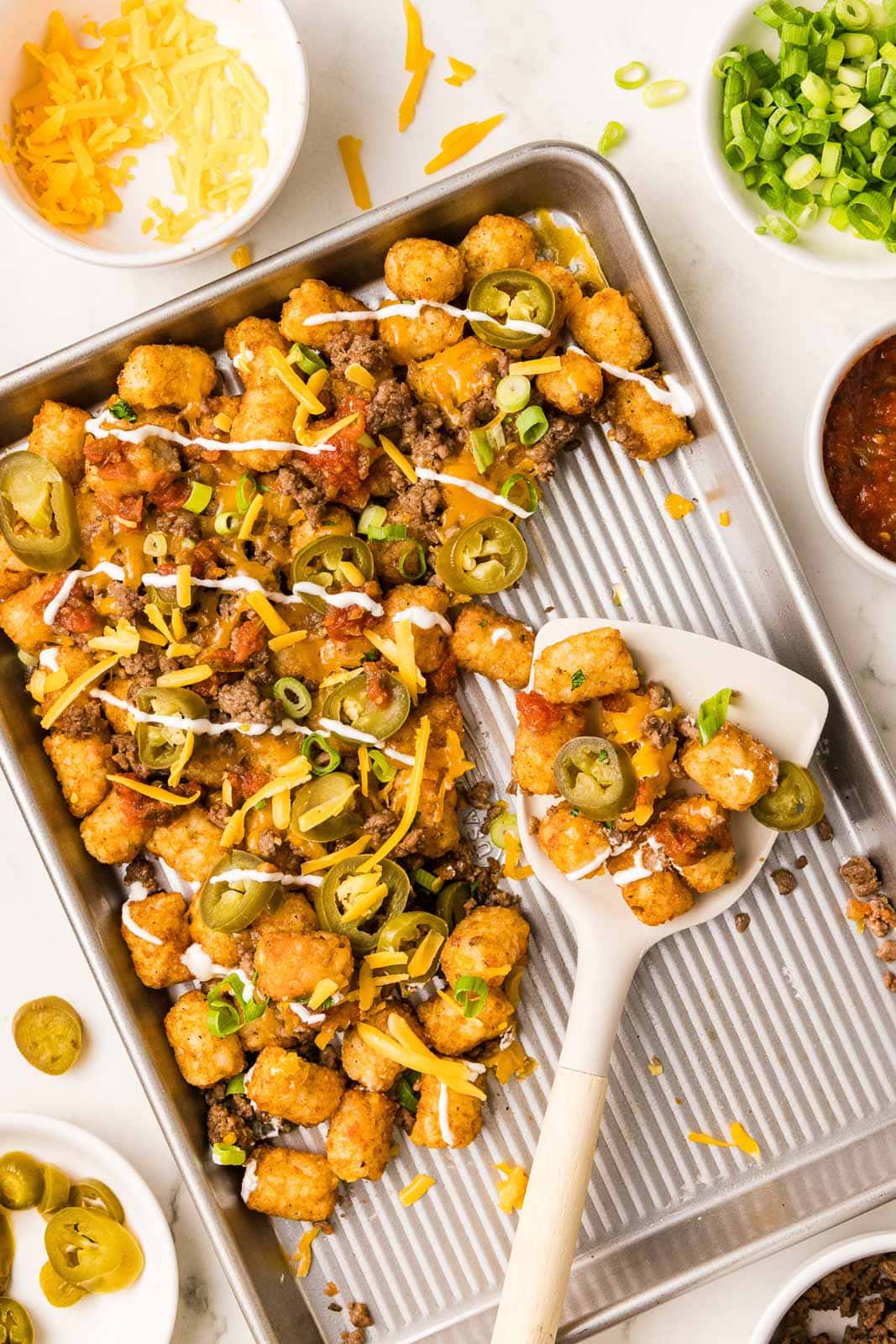 half-full sheet pan of tater tots with white spatula holding tater tots surrounded by bowls of cheese, green onion, salsa, and jalapeno