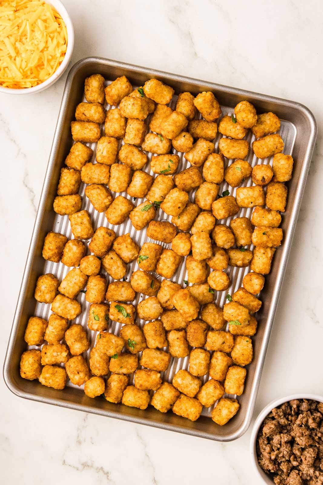 sheet pan containing tater tots with 2 small white bowls of ground beef and cheddar cheese