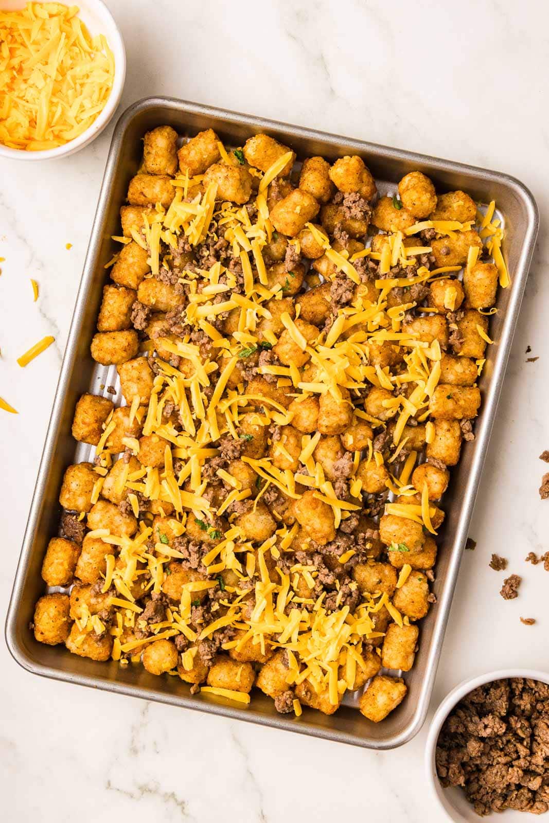 large sheet pain containing tater tots covered by ground beef and cheddar cheese, with 2 small bowls of ground beef and cheddar cheese