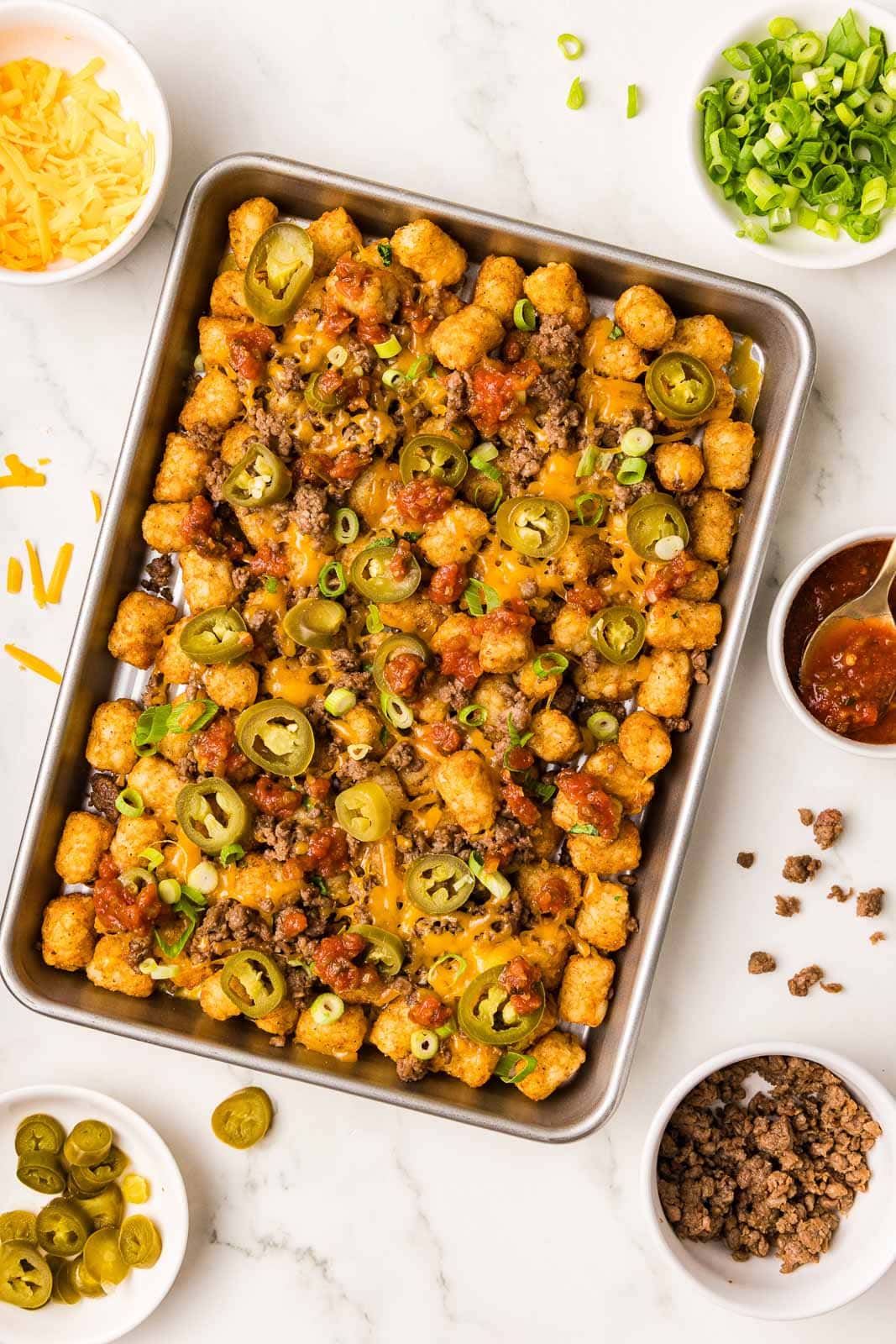 overhead view of completed tater tots on sheet pan with bowls of cheddar cheese, green onions, jalapeno, salsa and ground beef