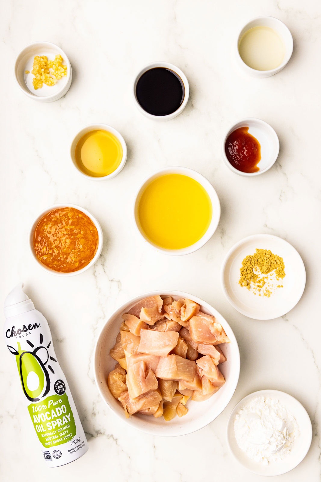 ingredients including white bowl with bite-sized chicken, can of avocado spray, along with small bowls of cornstarch, garlic, soy sauce, honey, hot chili sauce, orange marmalade, rice vinegar, orange juice and ginger