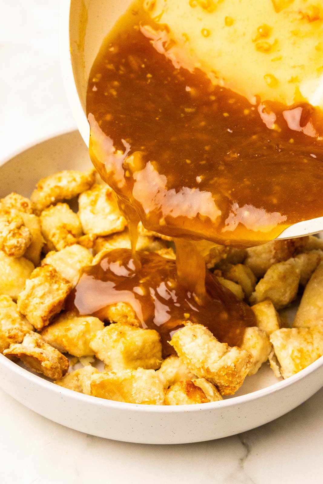closeup of orange mixture being poured out of skillet into white bowl containing air fried chicken pieces