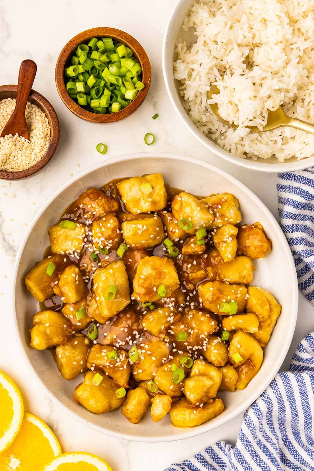 overhead view of prepared orange chicken pieces with topped with green onion and sesame seeds surrounded by white bowl of white rice, two wooden bowls with sesame seeds with wooden measuring spoons and green onions 3 lemon slices, and a blue and white striped dish cloth