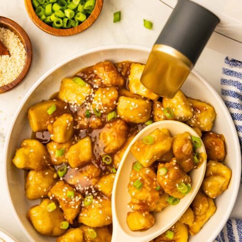 view of prepared orange chicken pieces topped with green onion and sesame seeds surrounded by white air fryer, two wooden bowls with sesame seeds with wooden measuring spoon and green onions, 3 lemon white bowl of prepared white rice, and a blue and white striped dish cloth