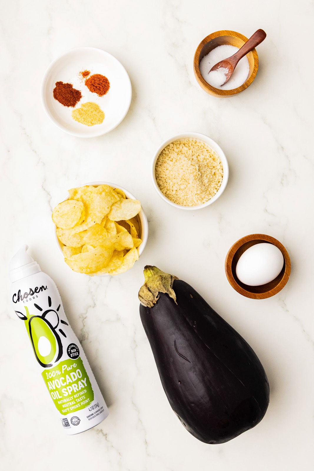 ingredients including eggplant, avocado oil spray, white bowls of potato chips, panko, seasonings, small wooden bowl of salt with spoon and one egg