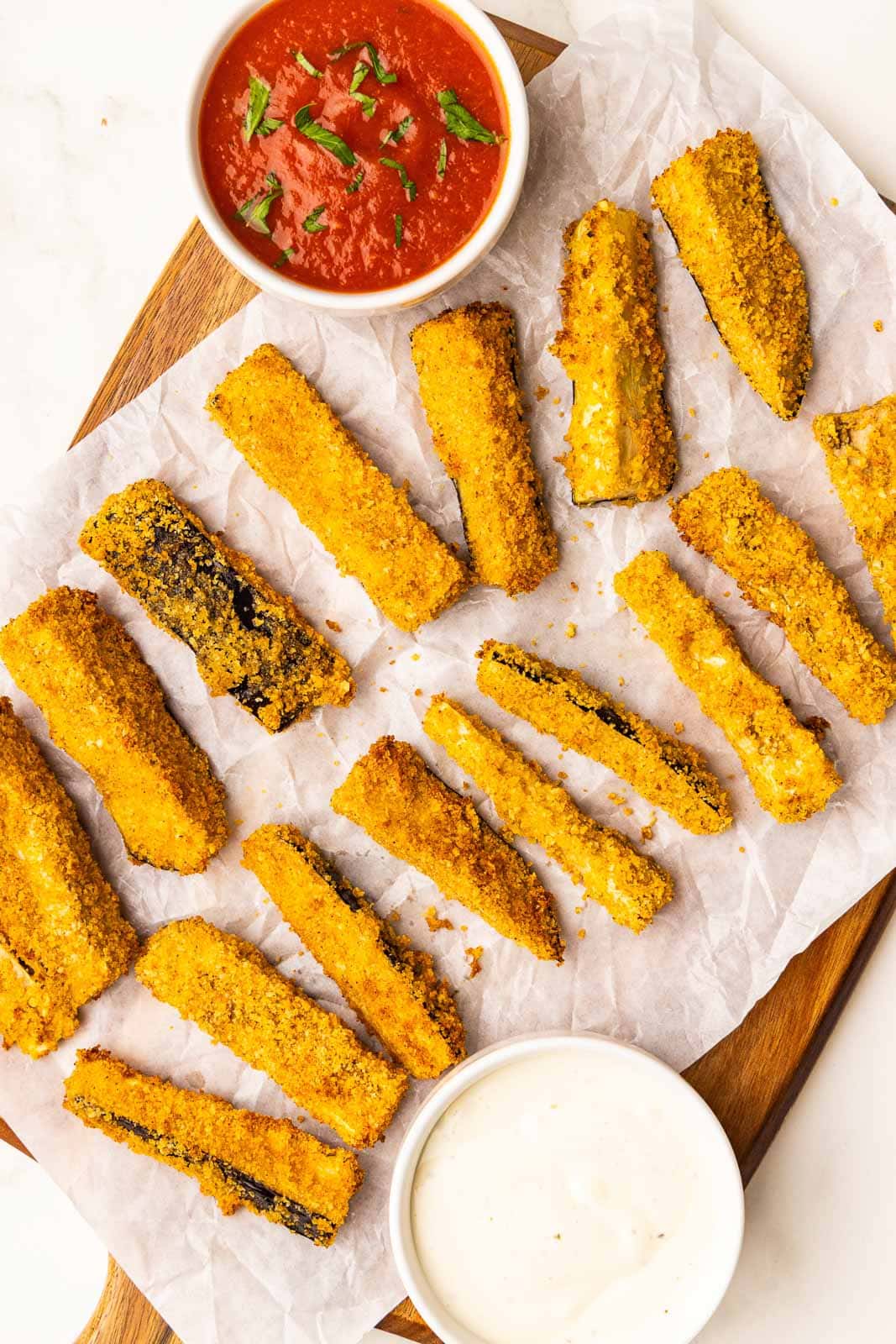 parchment lined wooden cutting board with 16 air fried eggplant sticks with white bowls of marinara and ranch dressing