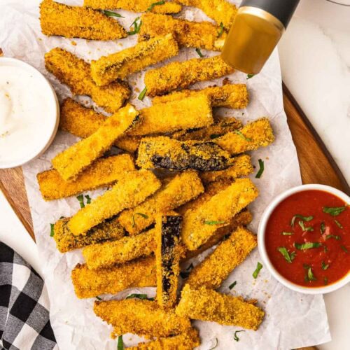 parchment lined wooden cutting board containing numerous eggplant sticks with bowls of marinara and ranch dressing alongside, black and white checked dish towel and white air fryer in background