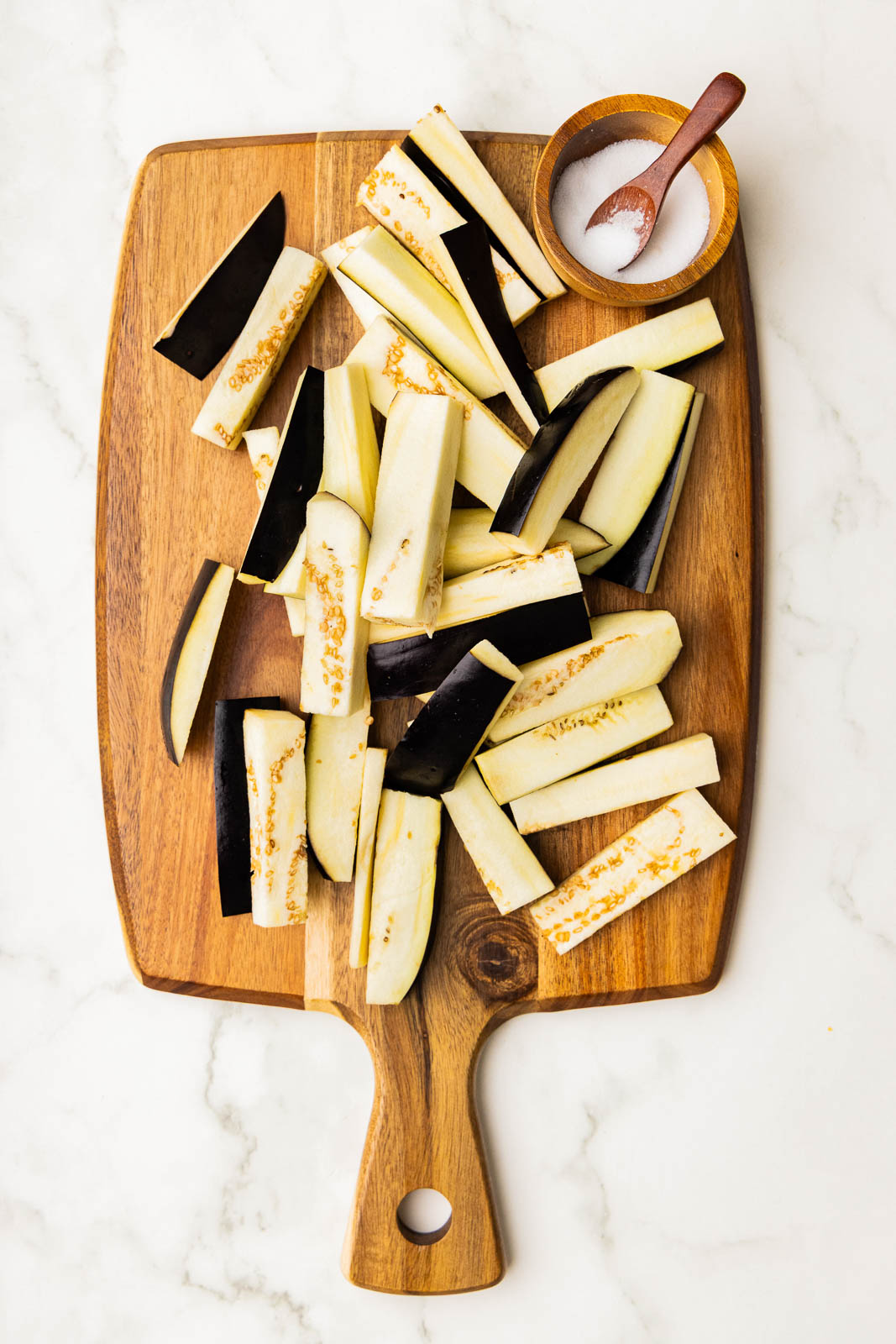 wooden cutting board with eggplant cut into sticks, with wooden bowl of salt and measuring spoon