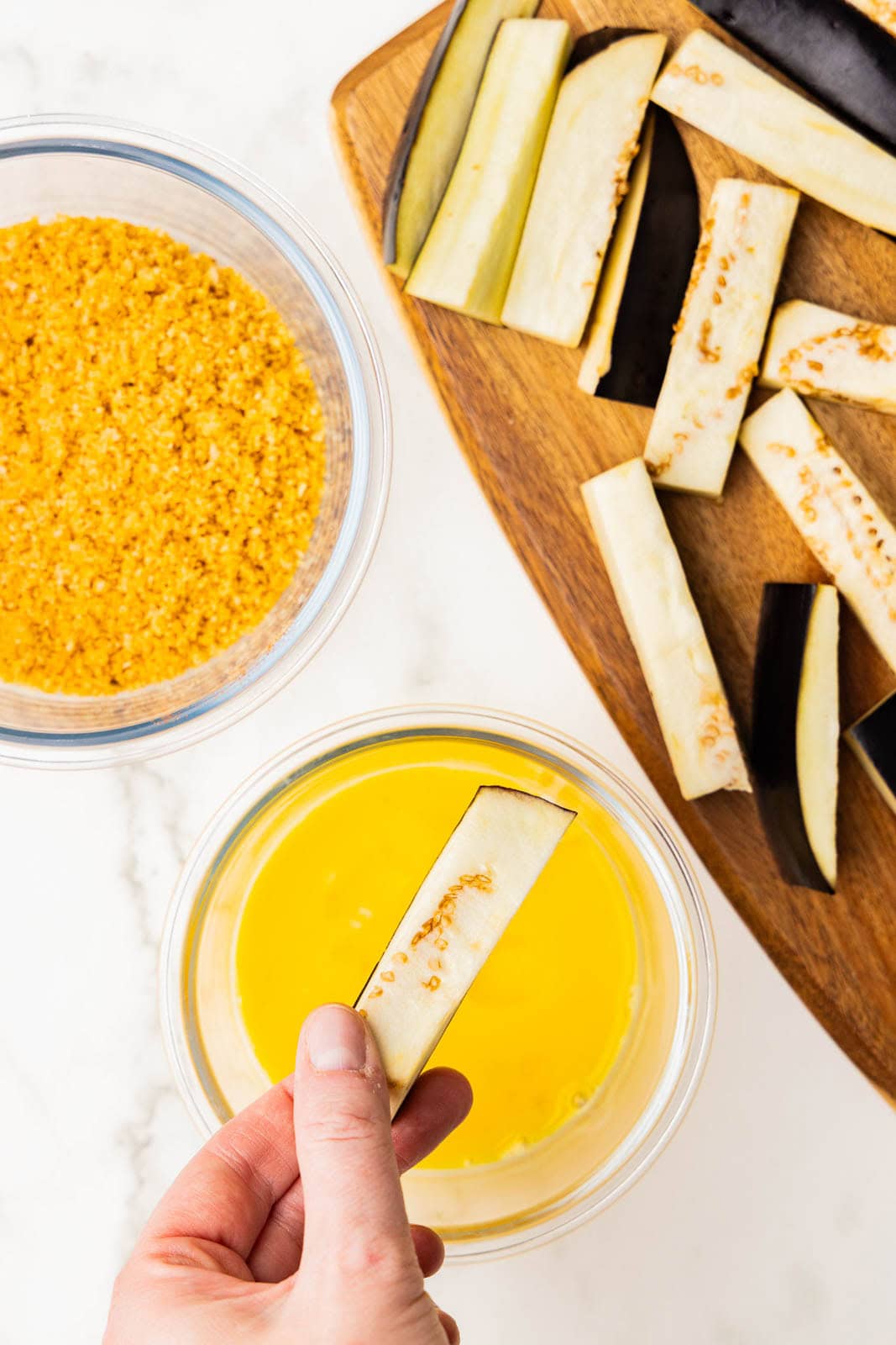 wooden cutting board with eggplant sticks, clear bowl of potato chips/panko crumbs and clear bowl of beaten egg with hand holding an eggplant stick