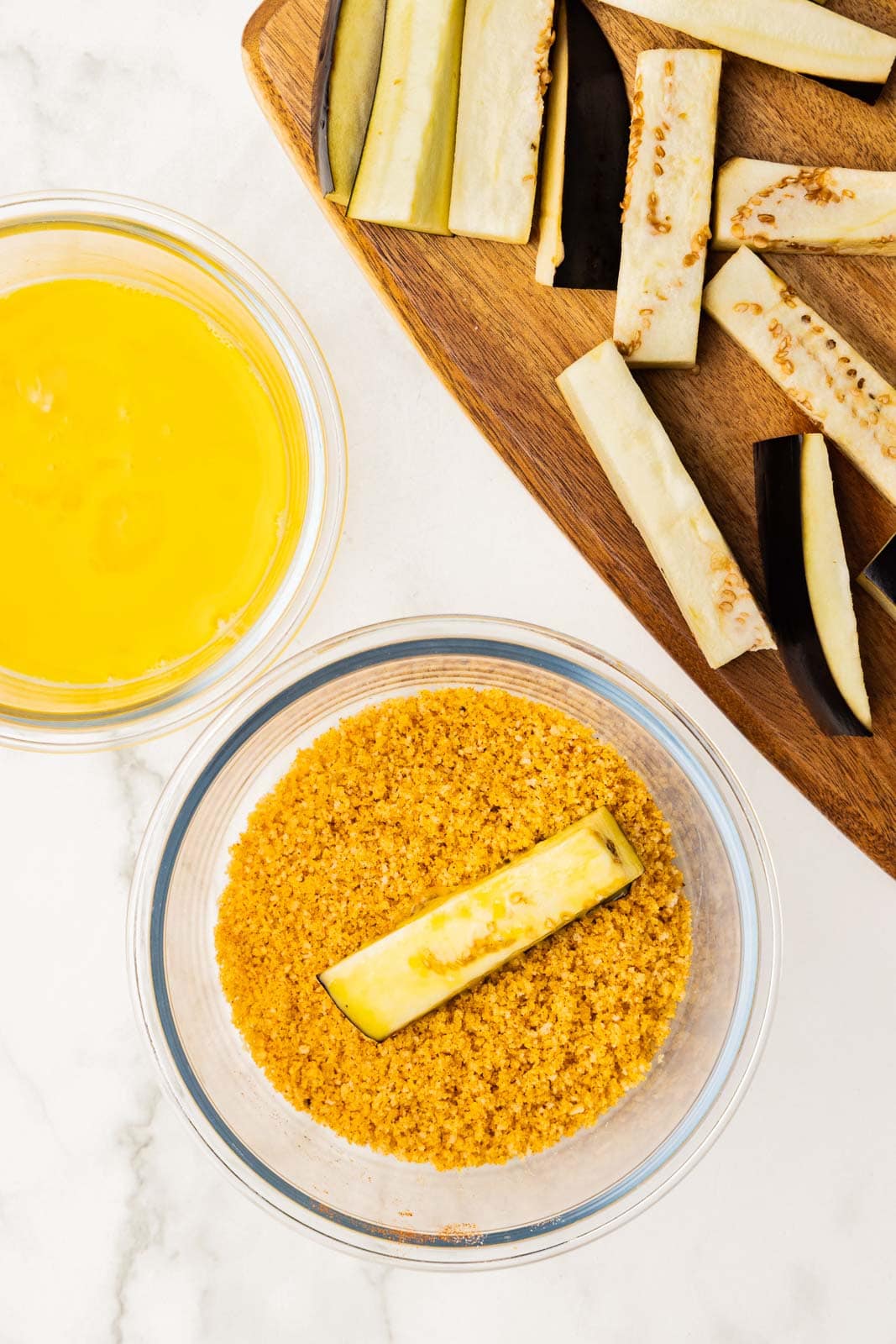 wooden cutting board with eggplant sticks, clear bowl of beaten egg and glass bowl of potato chips/panko crumbs with eggplant stick