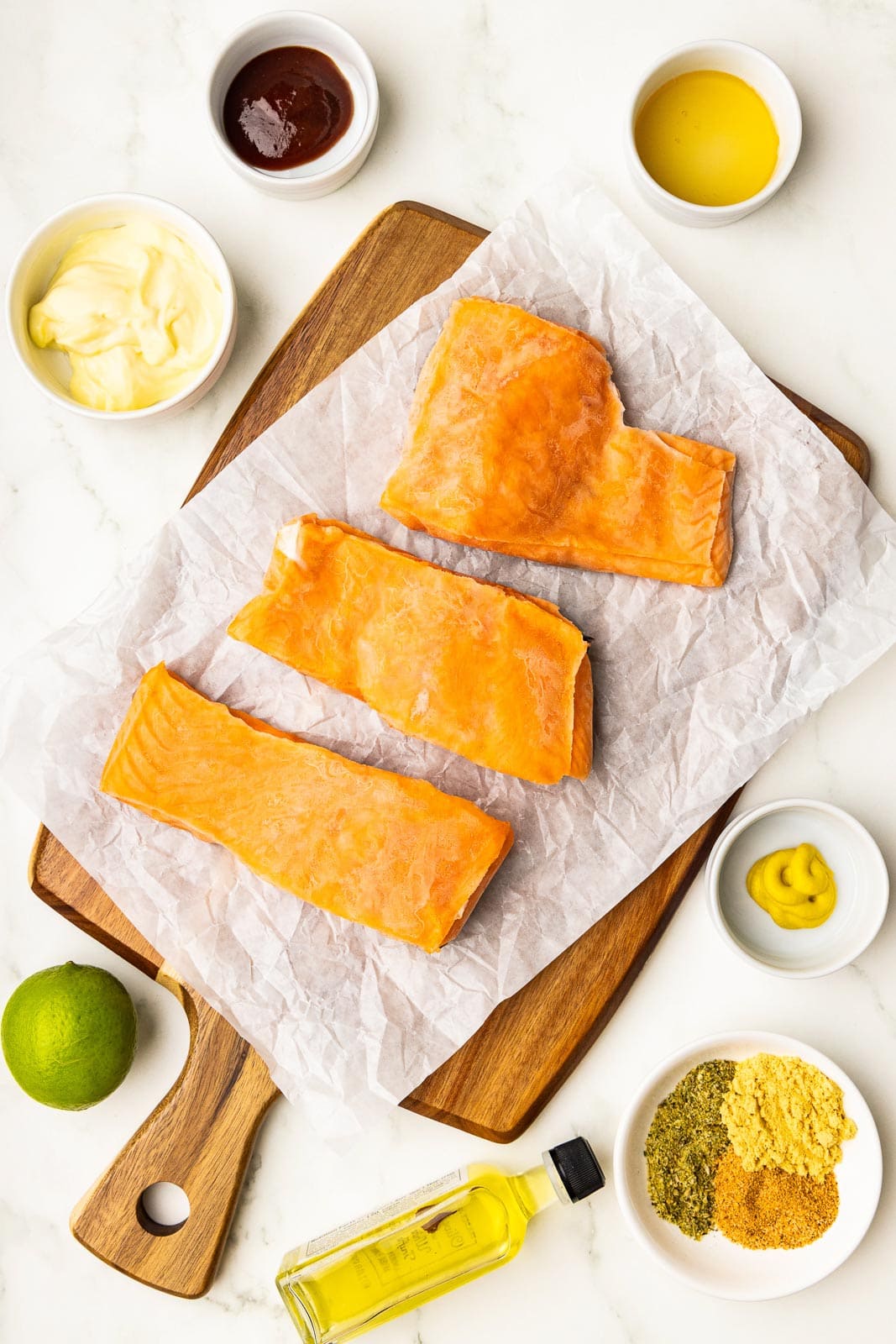 parchment lined wooden cutting board with 3 salmon fillets, surrounded by 1 lime, small bottle of olive oil, along with small white bowls of mayo, yellow and stone ground mustard, BBq sauce, seasonings and honey