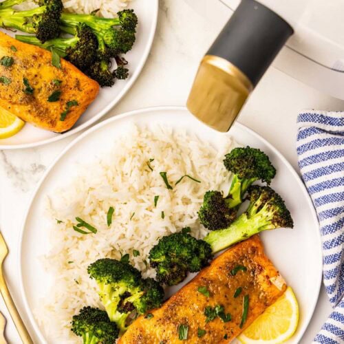 overhead view of 2 white dinner plates, each with 1 salmon fillet alongside white rice, broccoli and lemon slice, with 2 gold forks, blue and white striped dishcloth and white air fryer in background