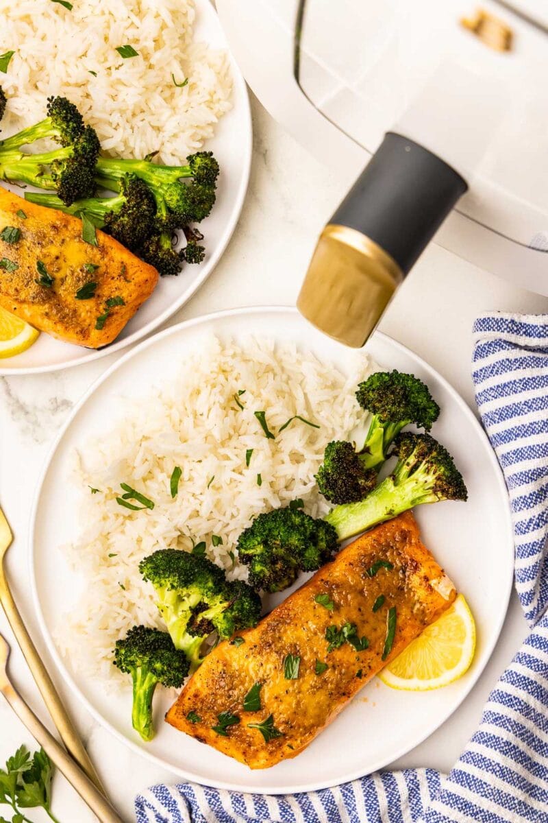 overhead view of 2 white dinner plates, each with 1 salmon fillet alongside white rice, broccoli and lemon slice, with 2 gold forks, blue and white striped dishcloth and white air fryer in background