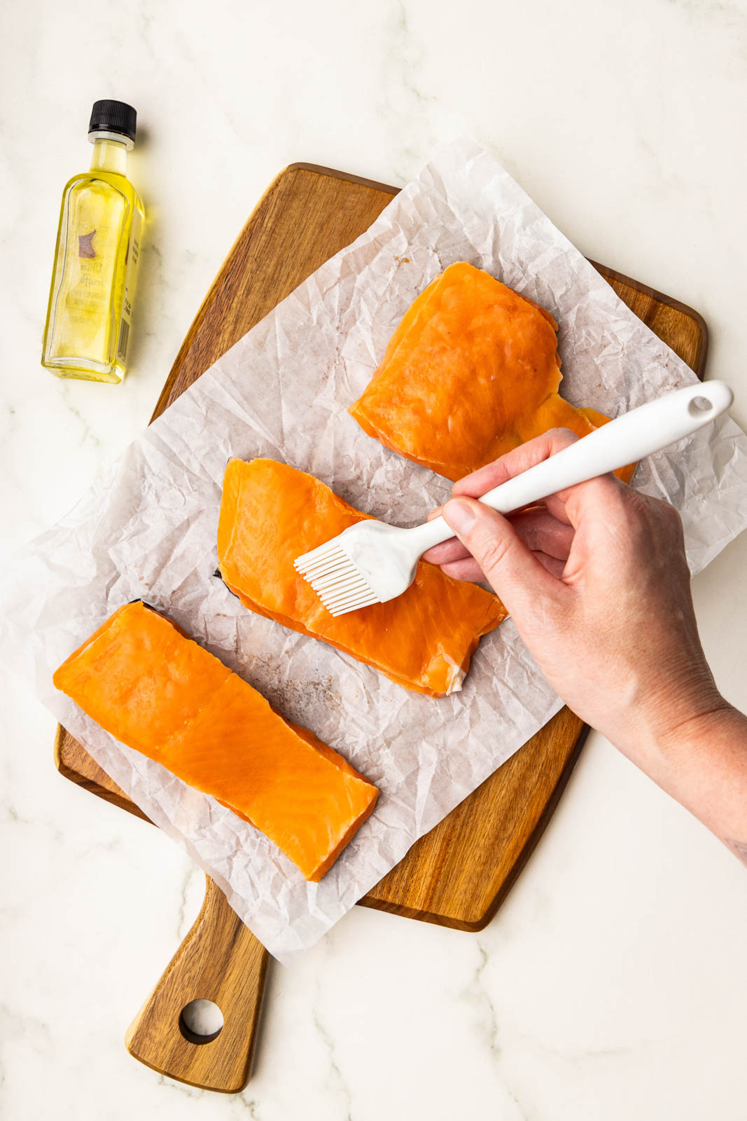 parchment lined wooden cutting board with 3 salmon fillets, small bottle of olive oil and hand busting one fillet with a white basting brush