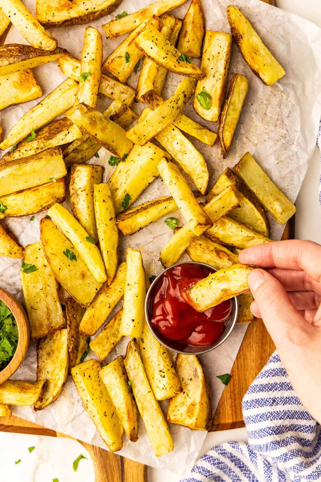 parchment paper lined wooden cutting board with air fried potato slices with handing dipping a slice into a bowl of ketchup