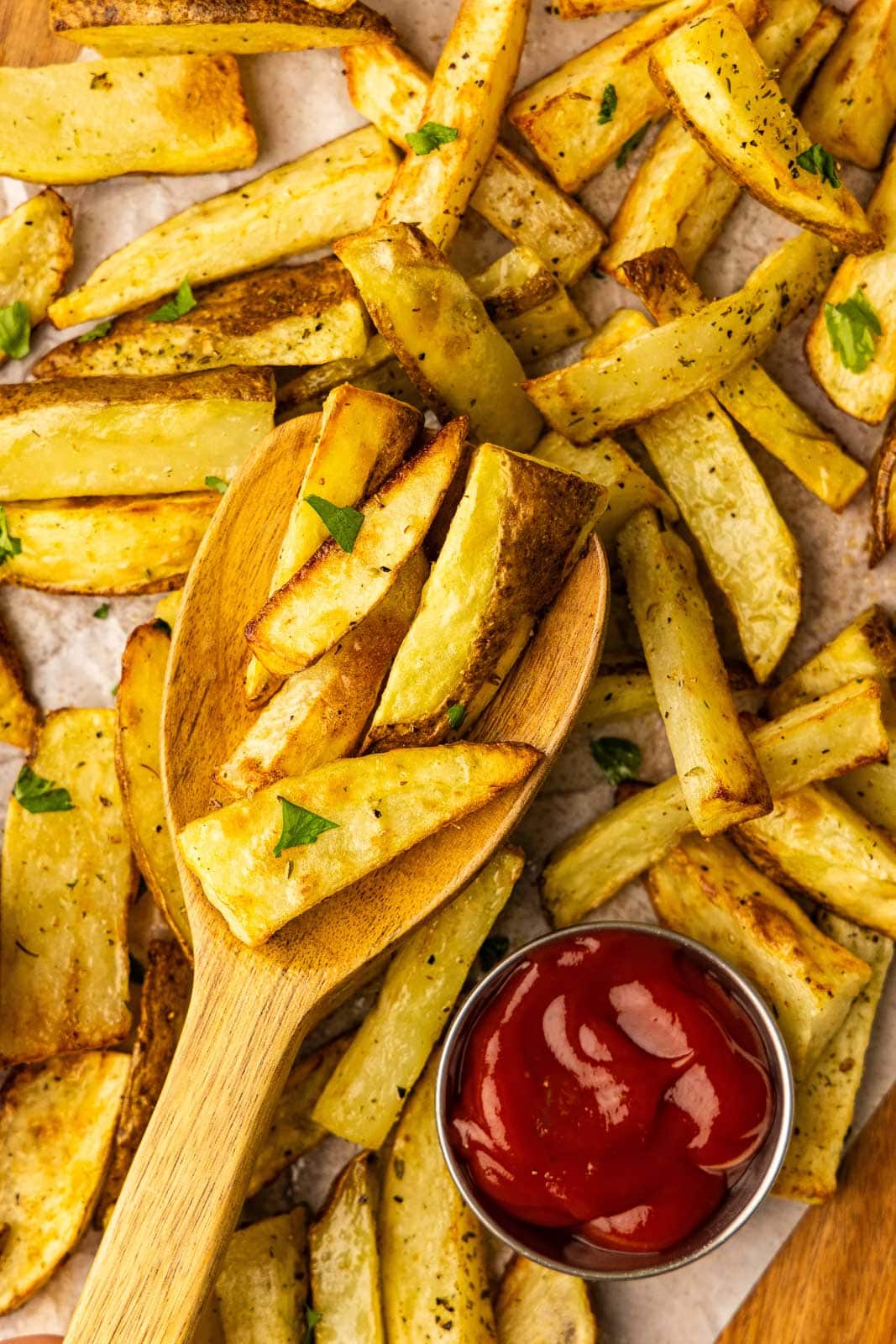 closeup view of parchment paper lined wooden cutting board of air fried potato slices with large wooden spoon and small bowl of ketchup