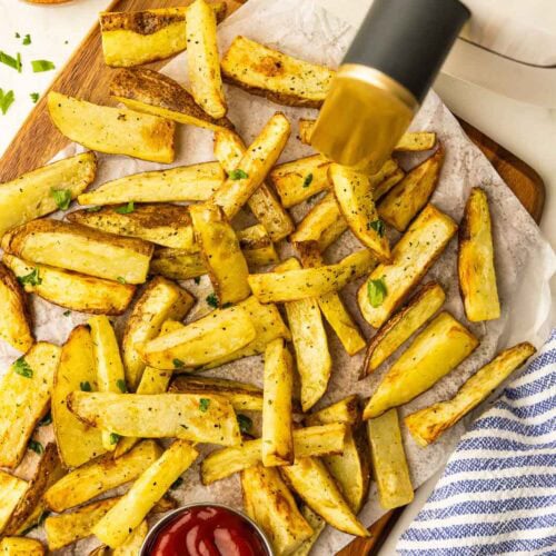 overhead view of parchment paper lined wooden cutting board containing air fried potato slices, small bowl of ketchup, with small wooden bowl of parsley and air fryer in background