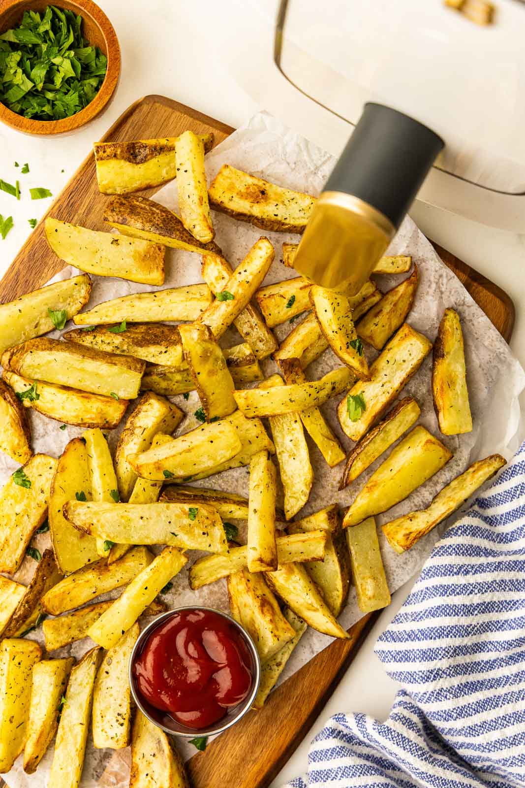 overhead view of parchment paper lined wooden cutting board containing air fried potato slices, small bowl of ketchup, with small wooden bowl of parsley and air fryer in background