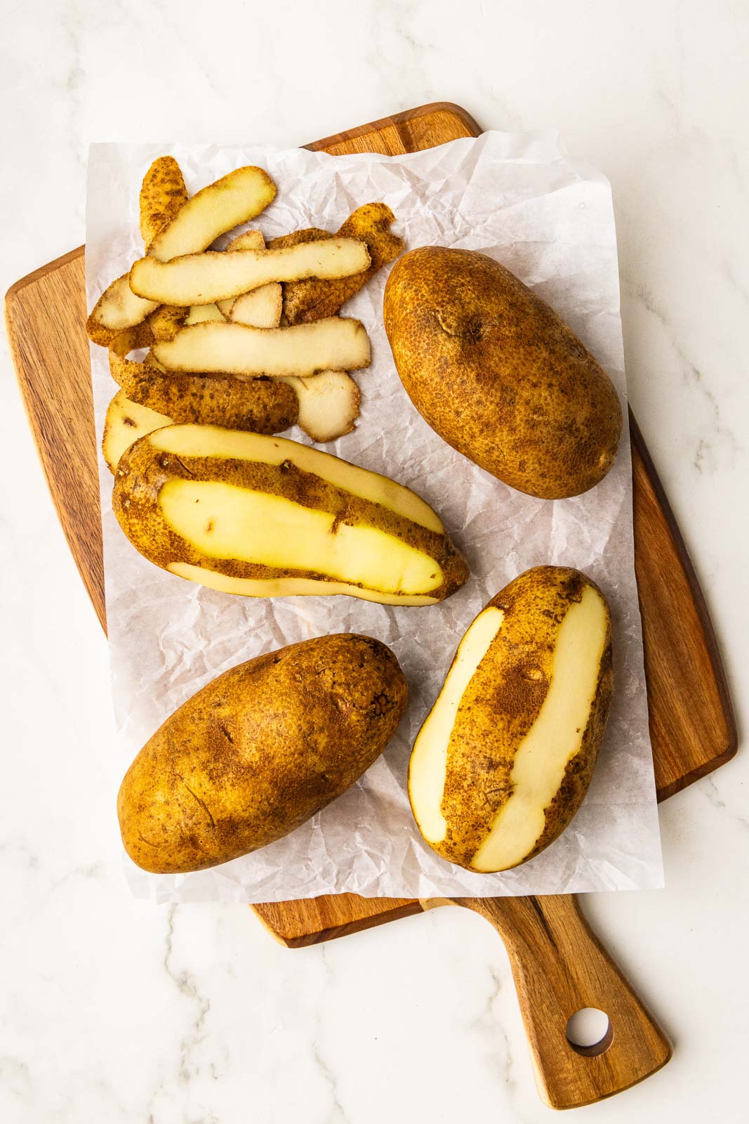 parchment paper lined wooden cutting board with 4 potatoes, 2 partially peeled with peelings
