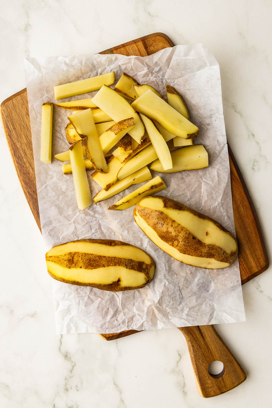 parchment paper lined wooden cutting board with 2 potatoes and partially peeled sliced potatoes