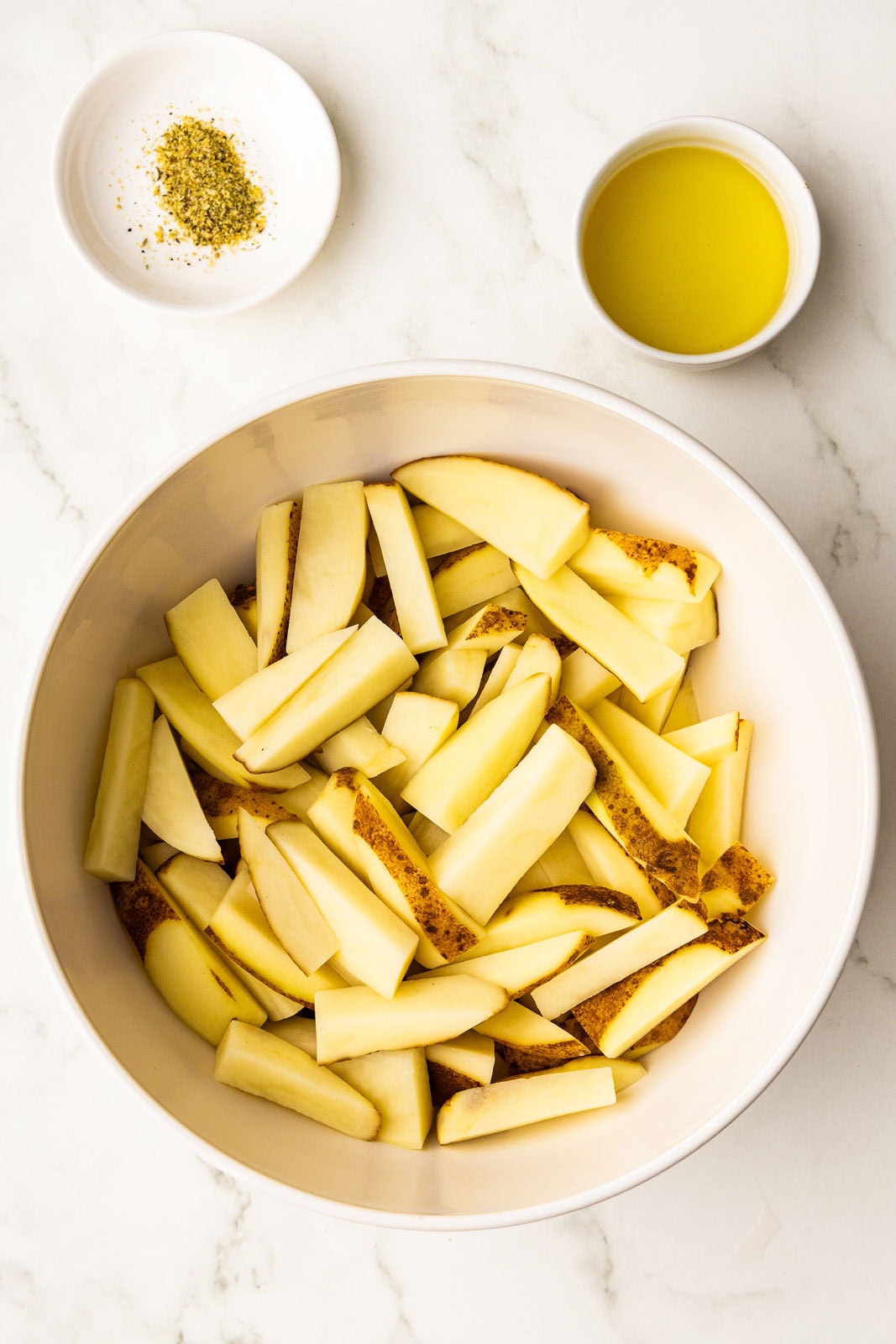 large white bowl containing sliced potatoes with 2 small bowls with avocado oil and seasoning