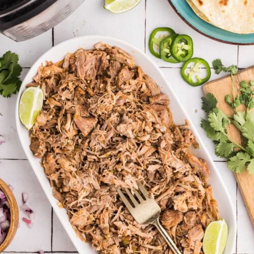 overhead view of white platter containing shredded pork, 2 lime wedges and gold fork, surrounded by instant pot, bowls of onions, cheese, plate of tortillas and cilantro