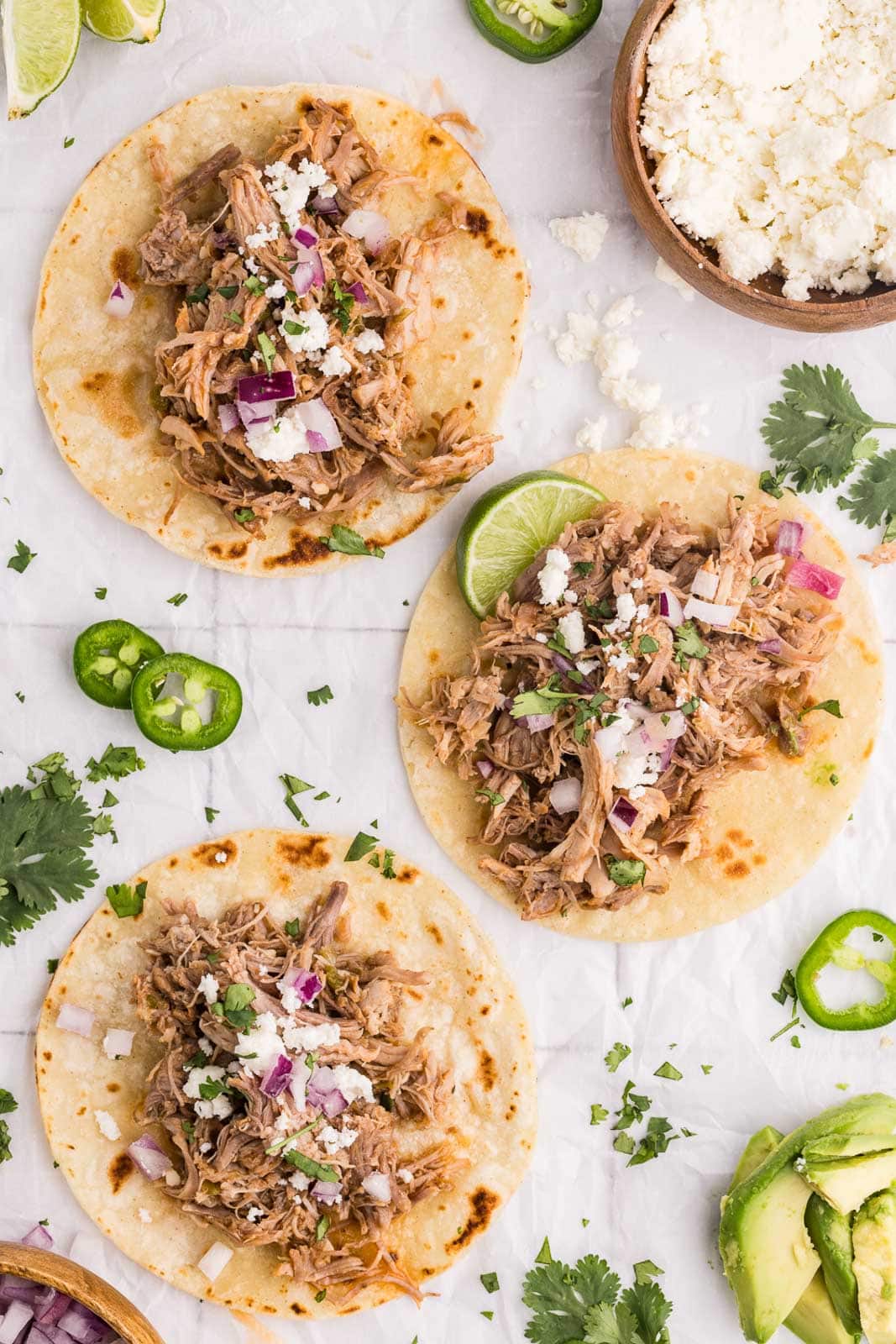 overhead view of three tortillas filled with pork mixture on parchment paper, with a bowl of cheese, jalapeno pepper slices, cilantro and avocado