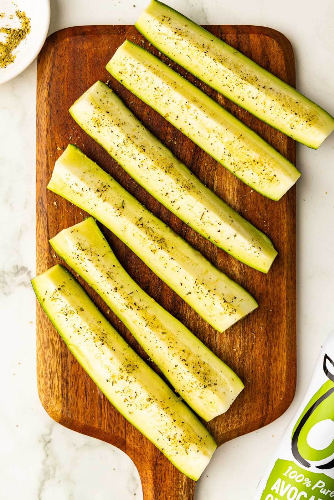 wooden cutting board with 6 slices of seasoned zucchini with spray and seasoning in the background
