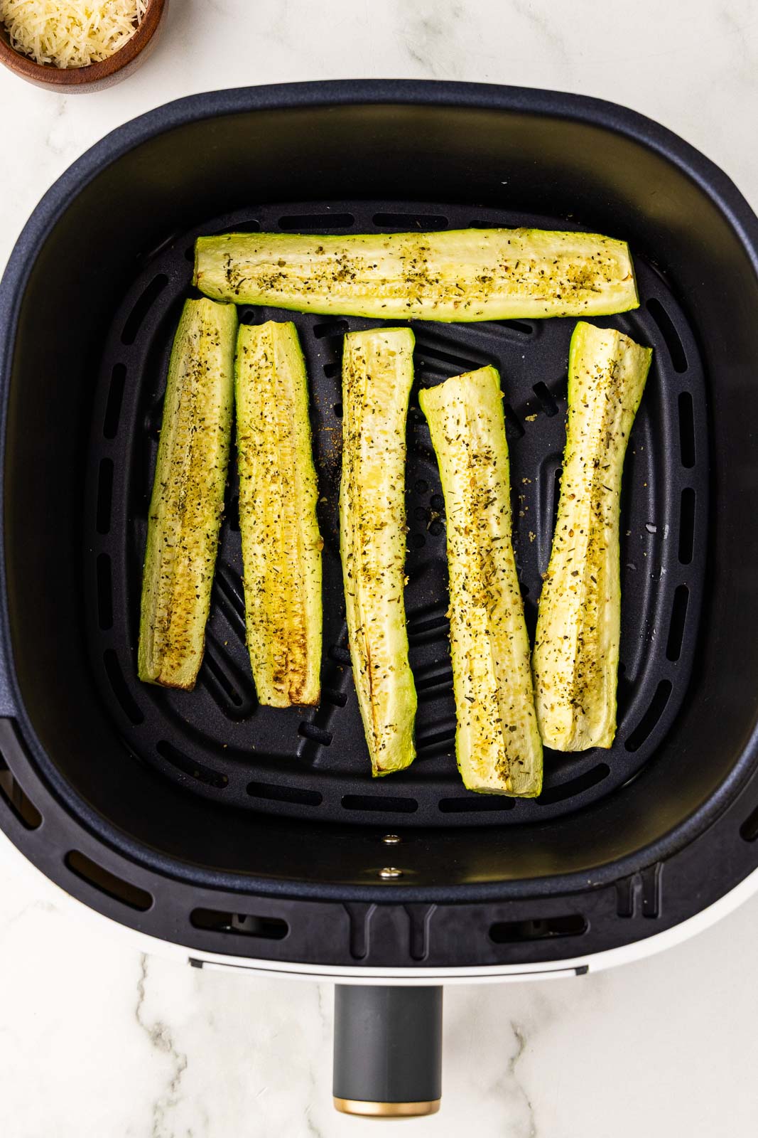 overhead view of black air fryer containing 6 air fried seasoned zucchini slices with bowl of Parmesan cheese in background