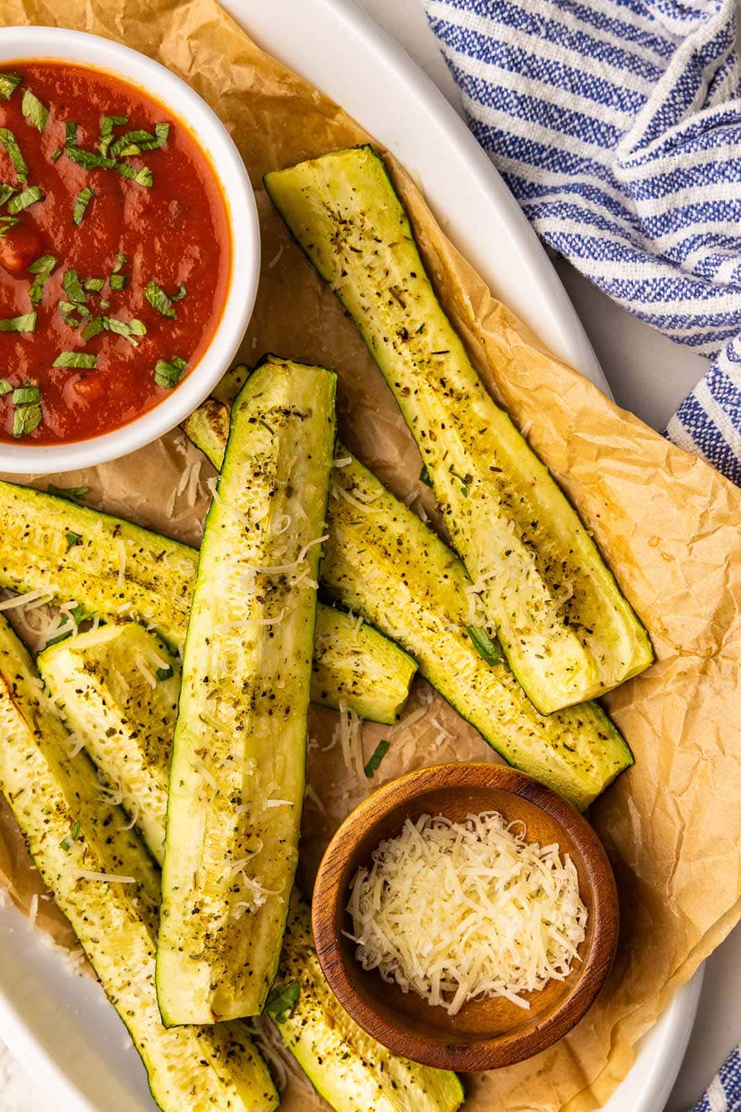 overhead view of parchment paper lined white serving platter with 6 slices of air fried zucchini topped with Parmesan cheese with small white bowl of marinara sauce and small wooden bowl of Parmesan cheese and blue and white striped dishcloth in background