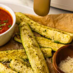 front view of parchment paper lined white serving platter with 6 slices of air fried zucchini topped with Parmesan cheese with small white bowl of marinara sauce and small wooden bowl of Parmesan cheese with air fryer in background