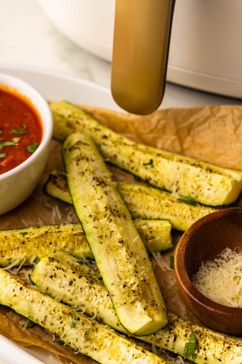 front view of parchment paper lined white serving platter with 6 slices of air fried zucchini topped with Parmesan cheese with small white bowl of marinara sauce and small wooden bowl of Parmesan cheese with air fryer in background