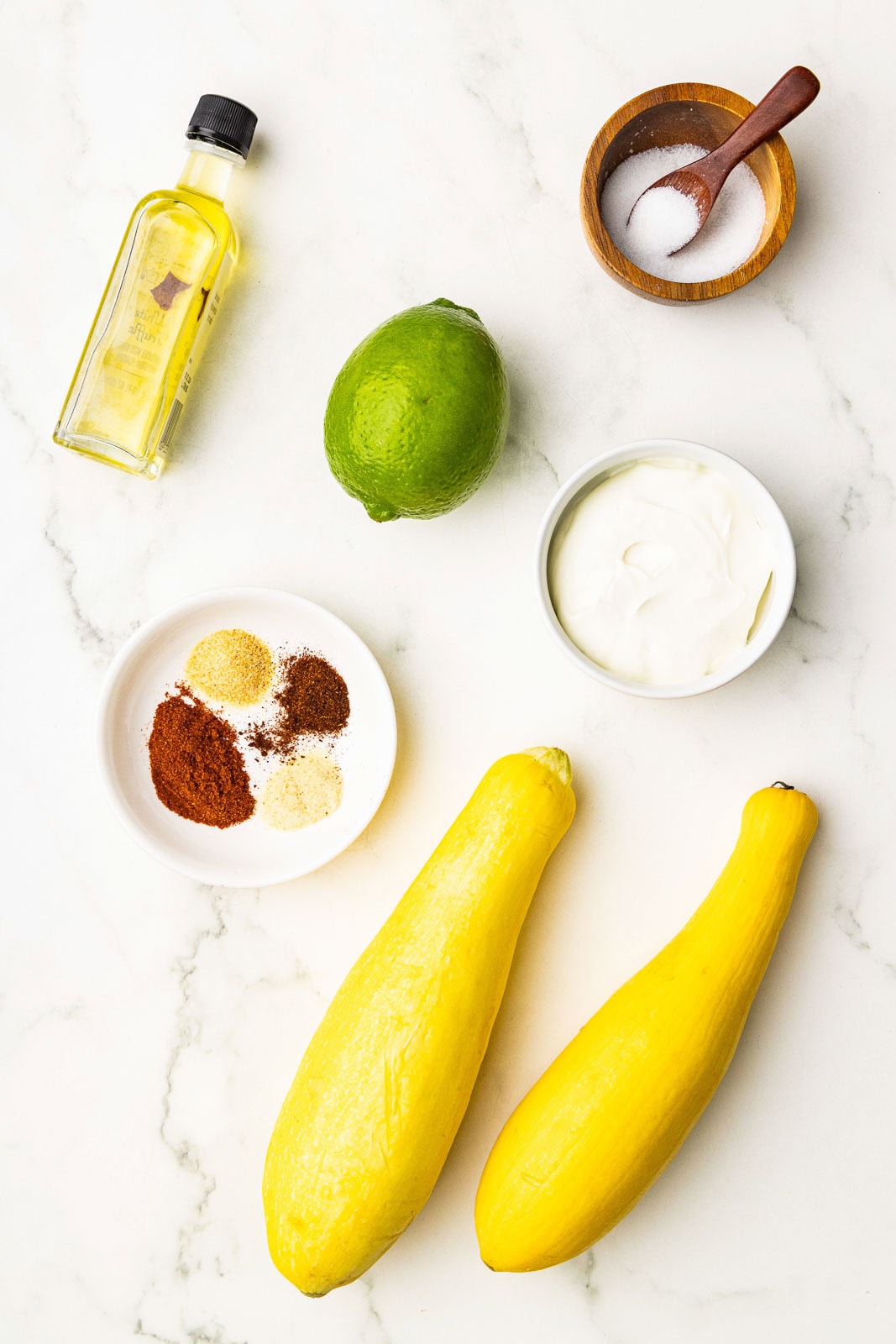 ingredients to include two yellow squash, bottle of olive oil, one lime, small white bowl of sour cream, wooden bowl and spoon of salt, and white bowl containing seasonings