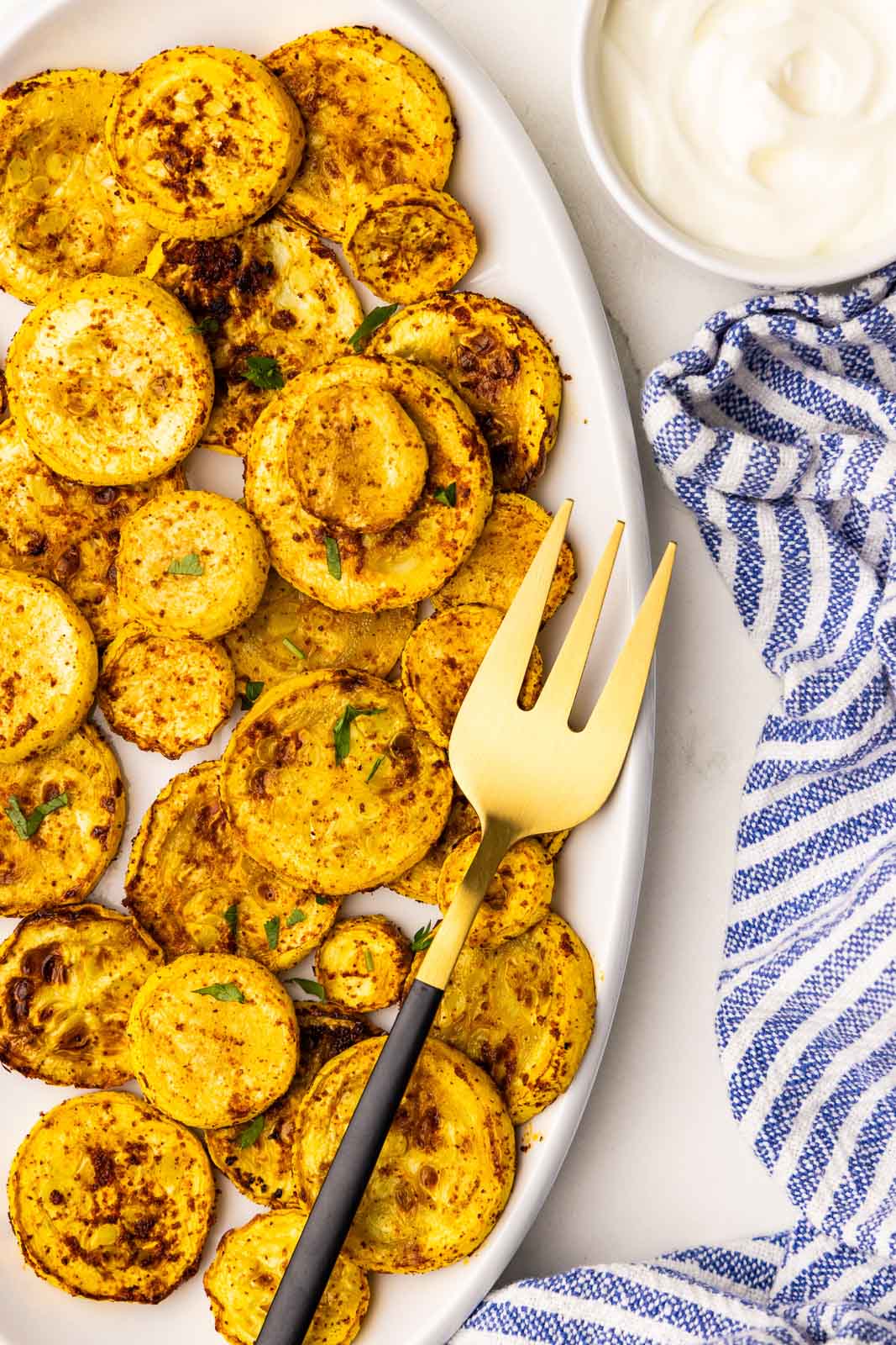 overhead view of white platter filled with air fried squash with gold fork, surrounded by small white bowl of sour cream and blue and white striped dish towel