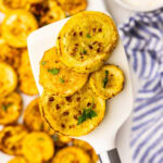 closeup of yellow squash on white spatula, with white platter filled with air fried squash with small white bowl of sour cream and blue and white striped dish towel