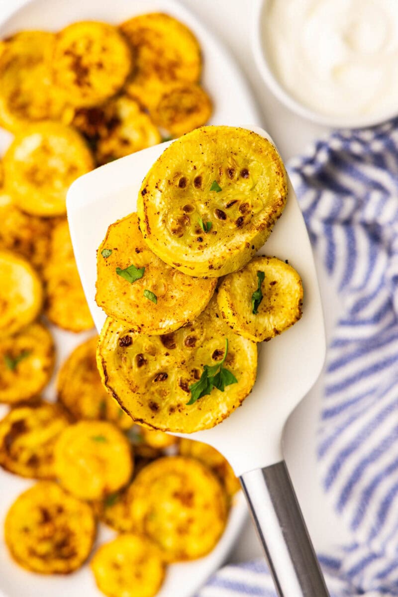 closeup of yellow squash on white spatula, with white platter filled with air fried squash with small white bowl of sour cream and blue and white striped dish towel