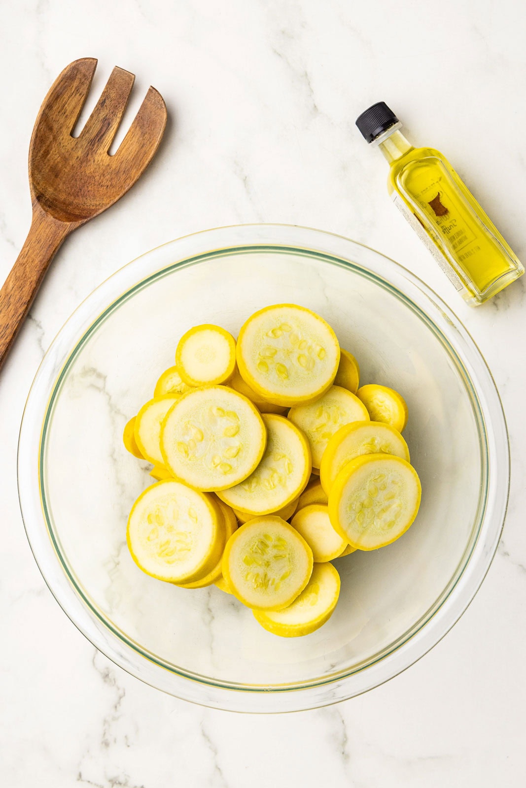 clear bowl containing sliced squash, surrounded by wooden fork and bottle of olive oil