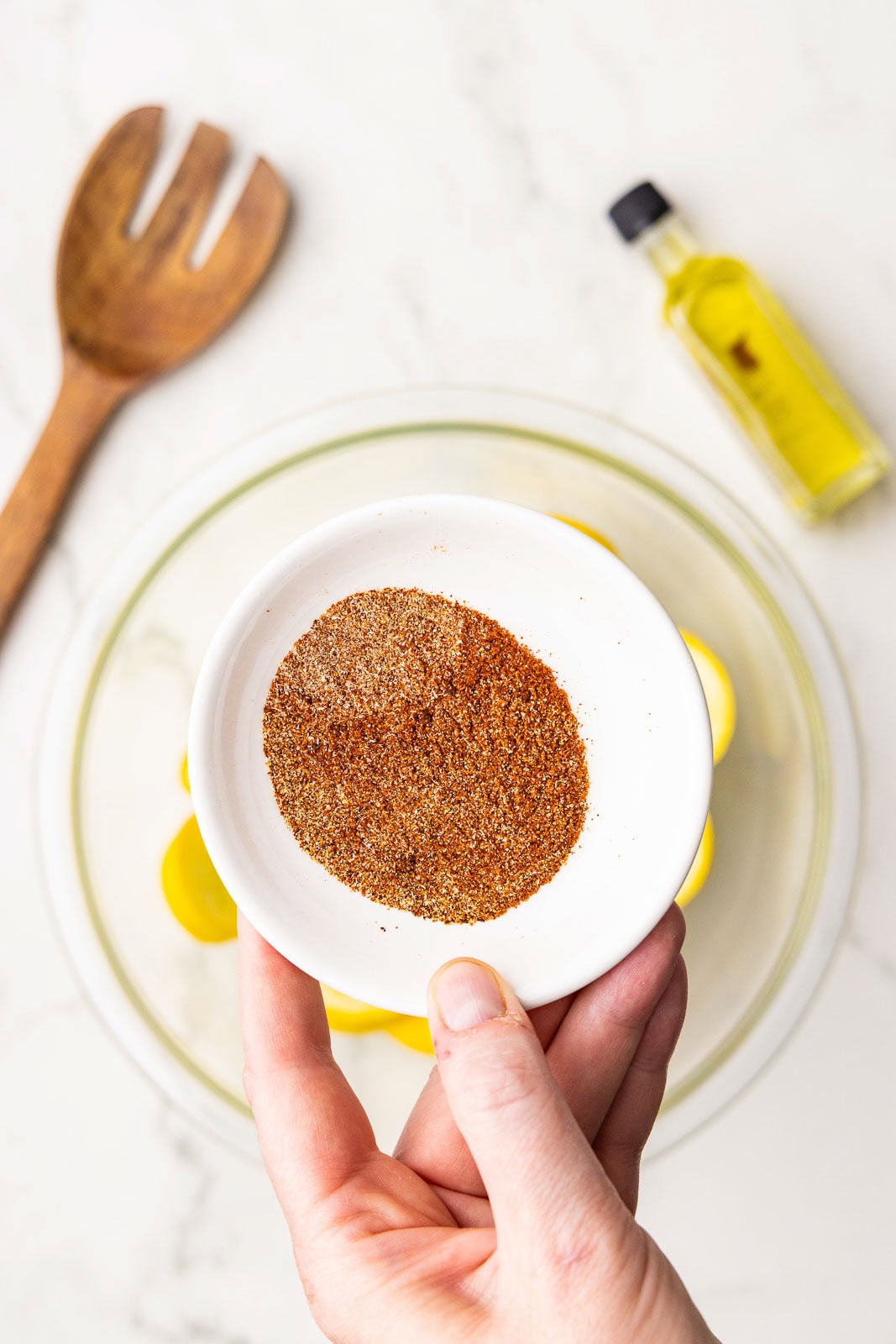 closeup of white bowl containing mixed seasonings, with clear bowl of sliced squash, wooden fork and bottle of olive oil in background