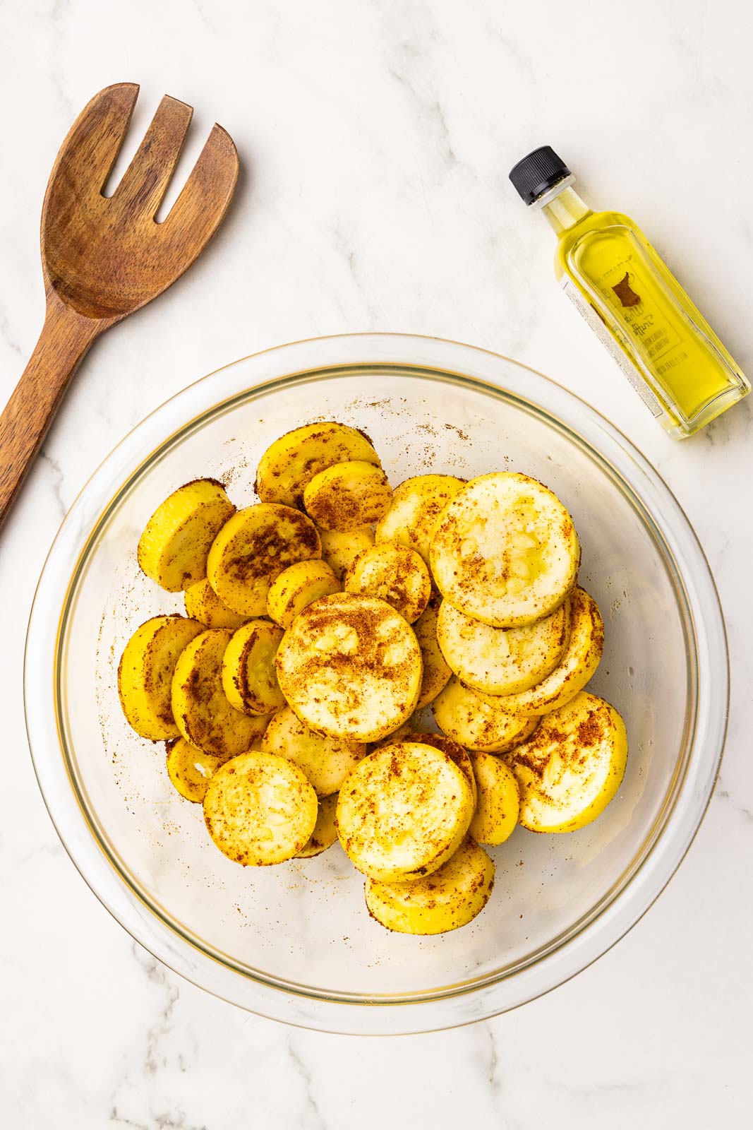 clear bowl containing seasoned sliced squash, surrounded by wooden fork and bottle of olive oil