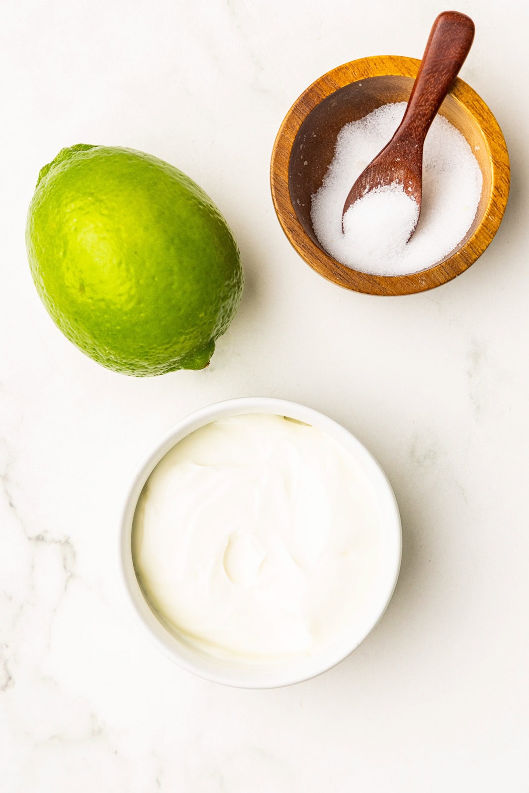 closeup of one lime, white bowl of sour cream and wooden bowl and spoon of salt