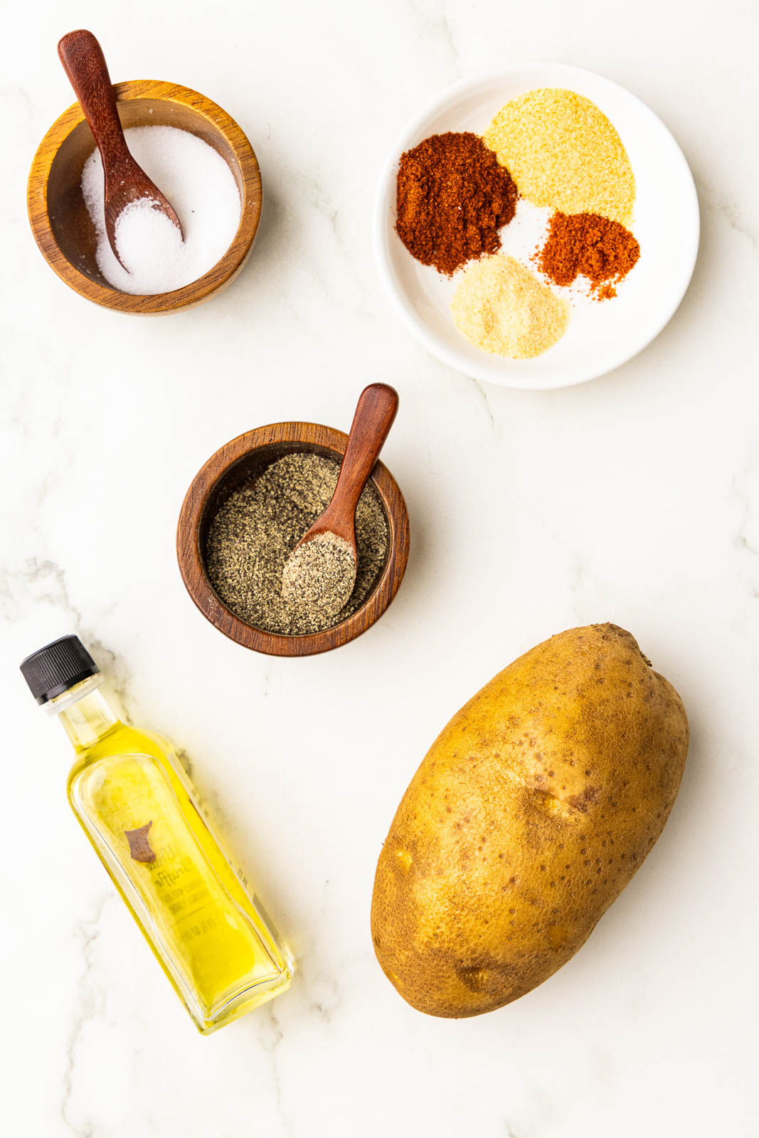 ingredients to include russet potato, bottle of olive oil, wooden bowls with spoons of salt and pepper, and white bowl of seasonings