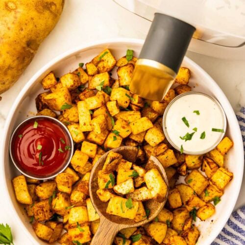 overhead view of white bowl containing air fried diced potatoes with containers of ketchup and ranch, surrounded by, parsley sprig, one uncooked potato, white air fryer, and blue striped dish towel
