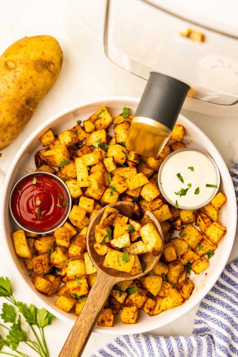 overhead view of white bowl containing air fried diced potatoes with containers of ketchup and ranch, surrounded by, parsley sprig, one uncooked potato, white air fryer, and blue striped dish towel