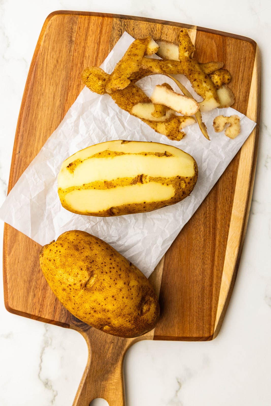parchment paper on wooden cutting board with one potato and one partially peeled potato with peelings on side