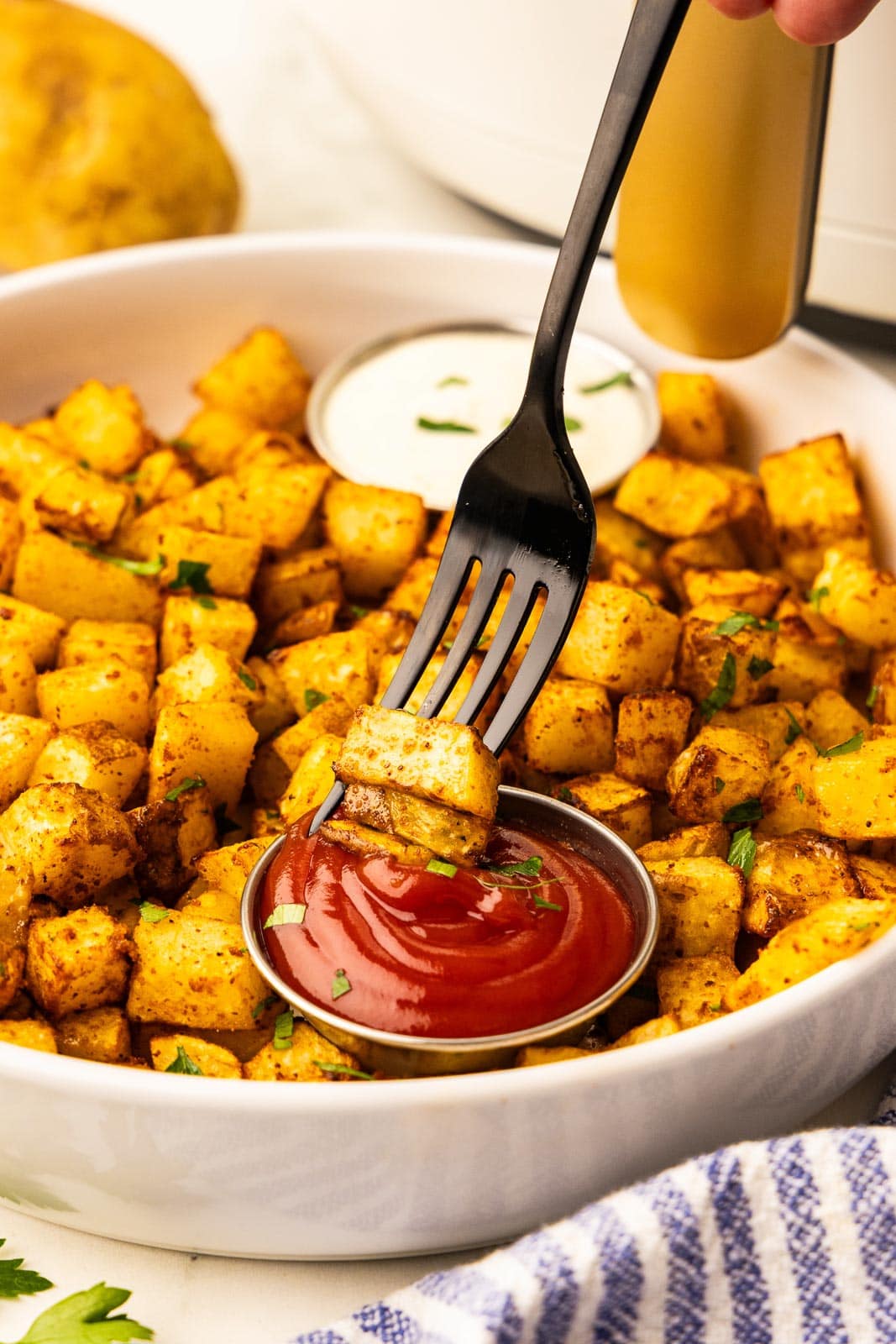 closeup of black fork with potato dipping into ketchup, with bowl of air fried potatoes and ranch dressing, with air fryer, potato and blue striped dish towel in background