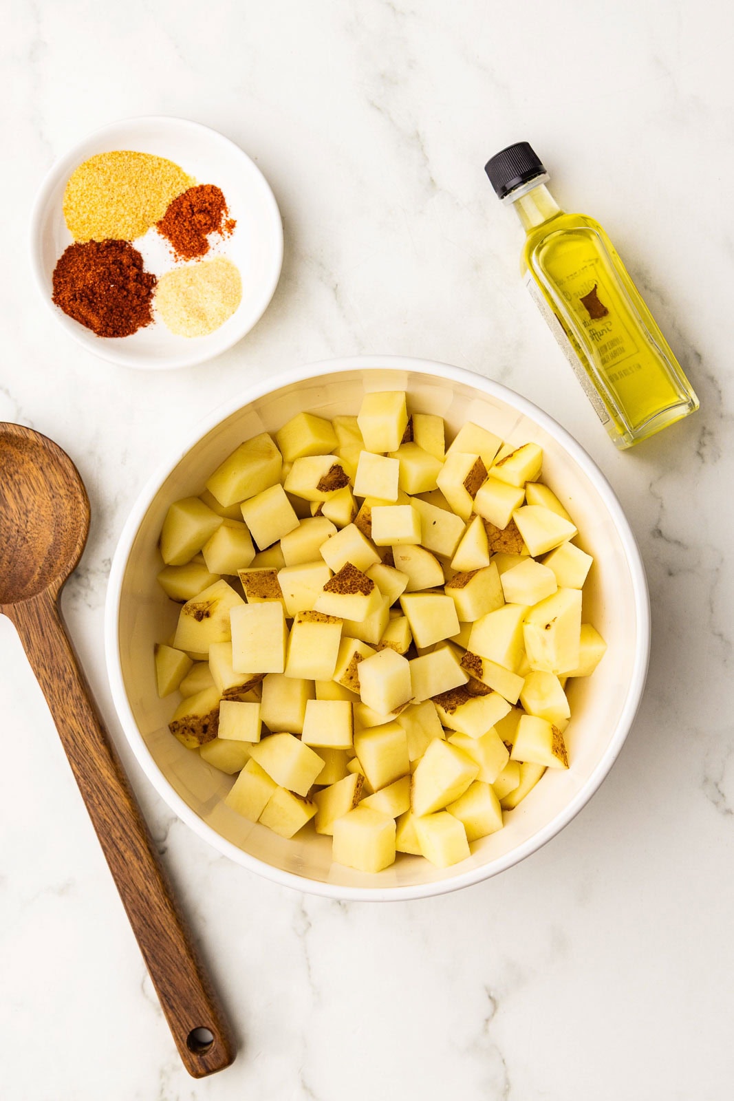 white bowl of diced potatoes, surrounded by wooden spoon, bottle of olive oil and small white bowl of seasonings