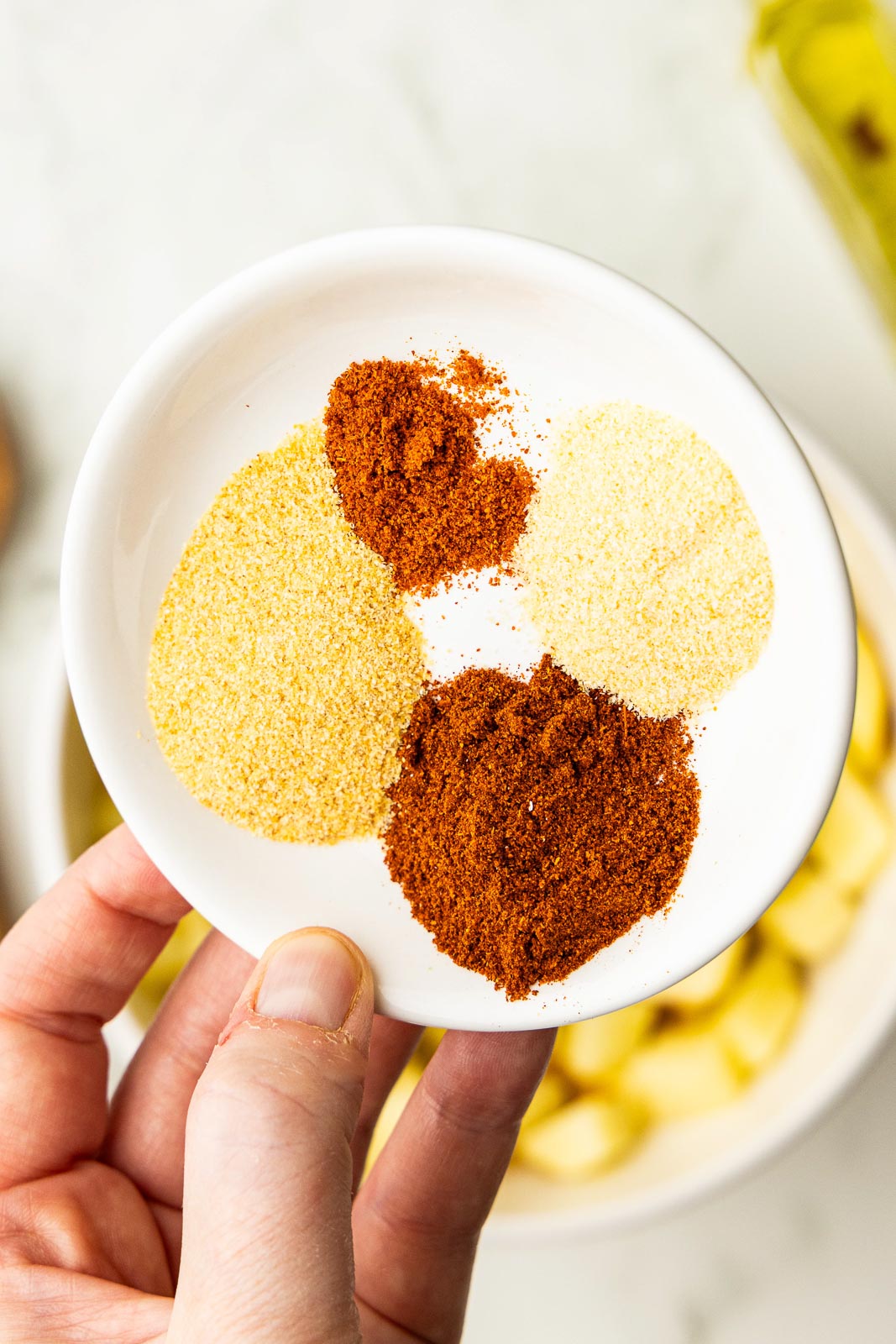 closeup of hand holding white bowl of seasonings with diced potatoes and olive oil bottle in background