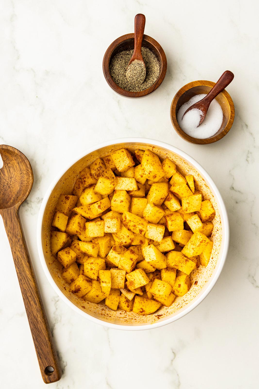 overhead view of white bowl containing seasoned diced potatoes, surrounded by wooden spoon and two wooden bowls with spoons containing salt and pepper